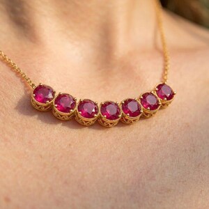 May include: A gold chain necklace featuring a curved row of seven round, faceted, deep pinkish-red gemstones. Each stone is set in a decorative, scalloped gold bezel setting, creating a continuous, elegant arc across the collarbone area.