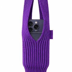 Könnte beinhalten: Eine leuchtend violette, gestrickte Handytasche mit gerippter Textur und einem langen, integrierten Griff. Die Tasche enthält ein dunkelgraues Smartphone mit einem Dreifach-Kamerasystem, das durch die Öffnung sichtbar ist.