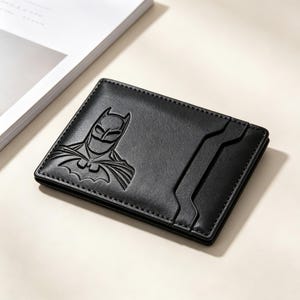 Puede incluir: Un tarjetero rectangular negro con el logotipo de Batman en relieve en la superficie frontal. La cartera está fabricada con un material liso e incluye un diseño de ranura vertical en el lado derecho para guardar tarjetas de forma segura.