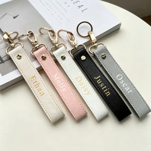 May include: Five rectangular faux leather keychains with gold-tone metal lobster clasps and rings. The straps are cream, pink, white, black, and gray, each featuring a gold or white embossed name: Ethan, Molly, Daisy, Justin, and Oscar.
