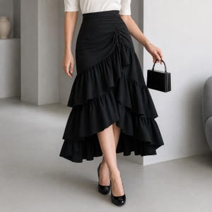 May include: A black high-waisted midi skirt featuring a ruched side detail with a drawstring tie, a tiered ruffled hemline, and an asymmetrical cut. The skirt is paired with a white short-sleeved top and a small black structured handbag.