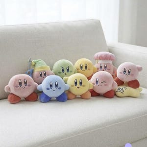 May include: A collection of nine round plush toys featuring the character Kirby in various colors and forms, including pink, blue, yellow, and green. Some figures wear accessories like a green hat or a chef hat, with one yellow base labeled 10th.
