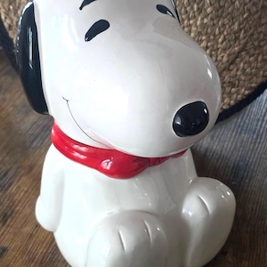 May include: A white ceramic figurine of a cartoon dog with black ears, a black nose, and closed eyes. The dog has a red scarf tied around its neck and is depicted in a seated position with its front paws resting on the ground.