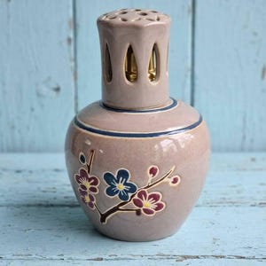 May include: A ceramic fragrance lamp with a glossy mauve finish, featuring a dark blue horizontal band near the top and a hand-painted floral branch design with blue, purple, and yellow blossoms. The lamp has a perforated metal crown.