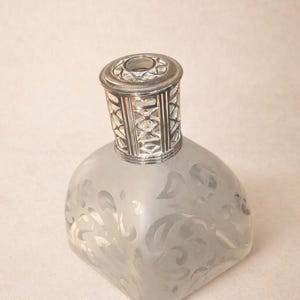 May include: A square-shaped fragrance lamp with a frosted glass body featuring a clear swirling scroll pattern. The top is capped with a metallic silver-toned openwork crown that has a geometric lattice design and a circular opening at the center.