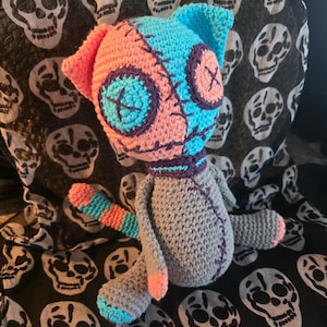 May include: A handmade crochet plush toy featuring a split-color head in bright pink and cyan, with dark purple button eyes stitched with an X pattern. The body is grey with purple stitched details, a striped tail, and pink and cyan accents on the limbs.