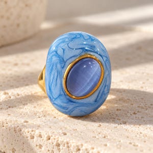 May include: A gold ring featuring a large, oval-shaped centerpiece with a light blue and white marbled enamel finish. The center of the piece contains a smooth, oval, periwinkle-colored cabochon stone set within a thin gold-toned bezel.