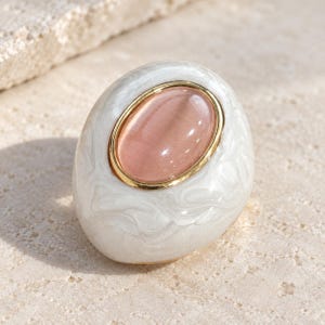 May include: A ring featuring a smooth, white, marbled-effect resin band with an oval, cabochon-cut, translucent pink stone set in a polished gold-tone metal bezel. The design is rounded and organic, highlighting the contrast between the pink stone.