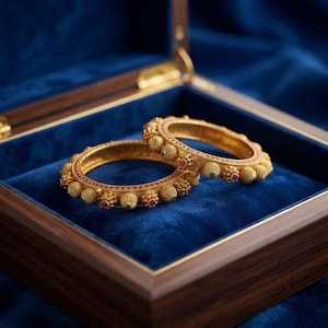 Puede incluir: Dos brazaletes dorados con una superficie texturizada y perlada, con pequeñas piedras rojas y verdes incrustadas. Las pulseras circulares presentan un patrón repetitivo de elementos esféricos en relieve a lo largo del borde exterior.