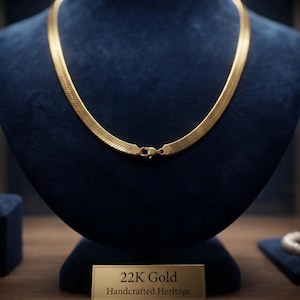 Puede incluir: Un collar de cadena de espiga en oro de 22 quilates con un diseño de eslabones planos y flexibles y un cierre de mosquetón seguro. El collar se exhibe sobre un busto de terciopelo azul oscuro con una placa rectangular que dice 22K Gold Handcrafted Heritage.