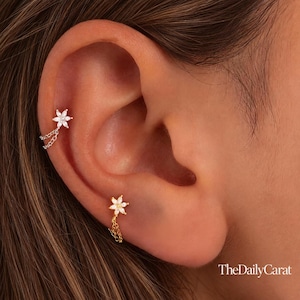 Puede incluir: Dos pendientes en forma de flor con cadenas colgantes. El pendiente superior es de tono plateado con piedras claras, y el inferior es de tono dorado con piedras claras. Ambos presentan un diseño floral de seis pétalos. Texto TheDailyCarat.