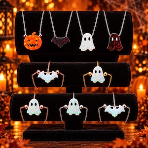 Op de afbeelding: Een collectie Halloween-sieraden op een zwarte standaard, bestaande uit vier kettingen met pompoen-, vleermuis- en spookhangers, en vijf armbanden met witte spook- en vleermuisbedels, allemaal voorzien van iriserende, kleurrijke details.