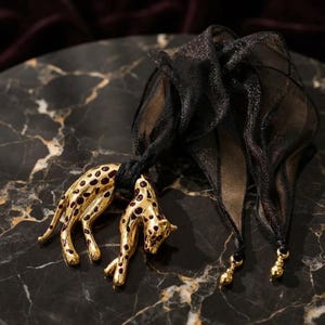 May include: A gold-toned metal brooch shaped like a leopard with dark red spots, attached to a sheer black fabric scarf. The scarf features a subtle shimmering texture and is finished with small gold-toned beads at the pointed ends of the fabric.