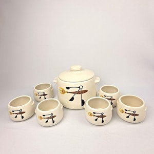 May include: A cream-colored ceramic serving set consisting of one lidded pot and six matching small bowls. Each piece features a repeating graphic design of a stylized brown and yellow fork and black ladle connected by thin black lines.