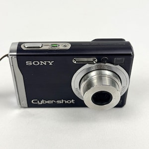 May include: A dark blue Sony Cyber-shot digital camera with a silver Carl Zeiss Vario-Tessar lens. The top panel features a silver power button with a green indicator light. The front displays the Sony and Cyber-shot logos in white text.