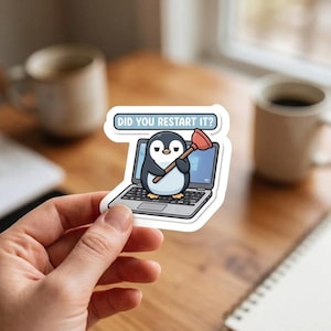 May include: A die-cut sticker featuring a cartoon penguin sitting on a laptop, holding a red plunger. Above the penguin, a speech bubble contains the text DID YOU RESTART IT? The sticker has a white border and a matte finish.