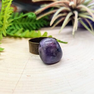 May include: A purple amethyst stone ring with a bronze band. The ring is on a light wood surface.