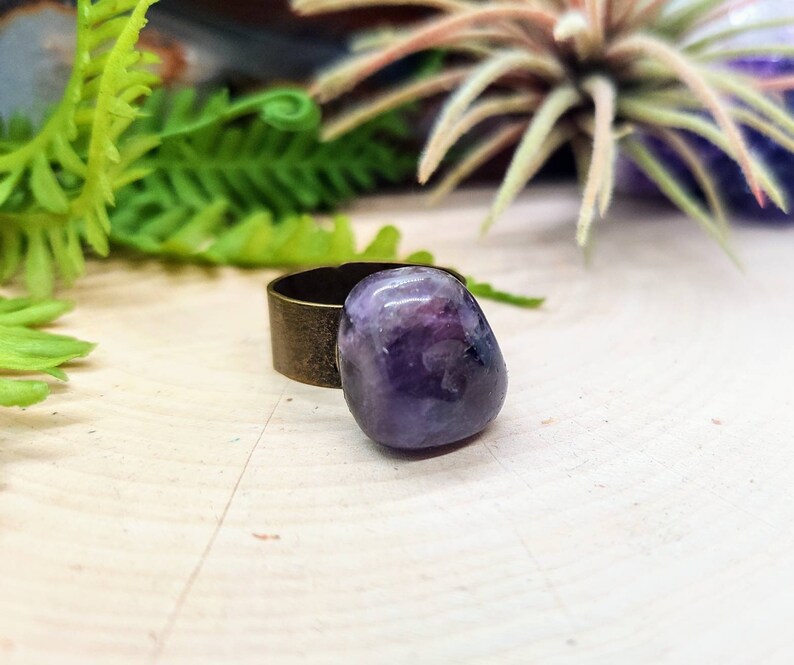 May include: A purple amethyst stone ring with a bronze band. The ring is on a light wood surface.