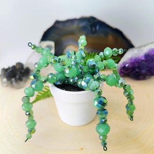 May include: A handcrafted beaded succulent plant in a small white pot. The plant features faceted green beads and iridescent accents, with wire stems. The pot sits on a wooden surface, with a geode and amethyst in the background.