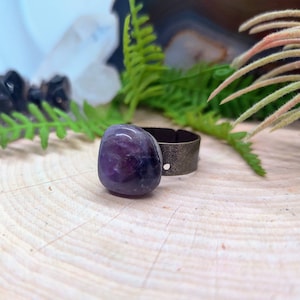 May include: A purple amethyst stone ring with a silver band. The ring is sitting on a wooden surface.