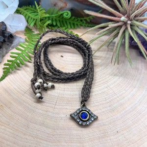 Op de afbeelding: Een bruin gevlochten koord ketting met witte kralen en een klein boze oog hanger met een blauwe steen en heldere strass steentjes.