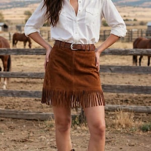 May include: A white long-sleeved button-down shirt paired with a brown suede mini skirt featuring a fringe hem and a brown leather belt with a silver buckle, complemented by brown leather cowboy boots with intricate stitching details.