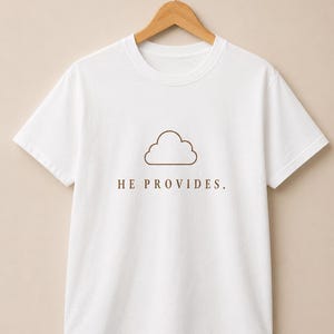 May include: A white short-sleeved t-shirt featuring a brown outline of a cloud graphic centered on the chest. Below the cloud, the text HE PROVIDES. is printed in a brown, capitalized, serif font. The shirt is displayed on a wooden hanger.