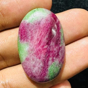 Puede incluir: Una piedra preciosa en forma de cabujón ovalado que presenta una mezcla vibrante de inclusiones minerales de color rosa magenta intenso y verde lima brillante. La superficie es pulida y lisa, mostrando una textura cristalina natural.