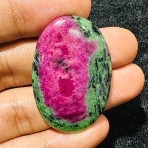 Puede incluir: Una piedra preciosa en forma de cabujón ovalado con un centro rosa magenta vibrante rodeado por una matriz de zoisita verde y negra moteada. La superficie pulida muestra un acabado liso y brillante con variaciones de color distintivas.