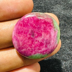 Op de afbeelding: Een ronde, gepolijste edelsteen met een levendig magenta-roze centrum en gevlekte groene randen. De steen heeft een glad oppervlak in cabochon-stijl met zichtbare natuurlijke mineraalinsluitingen en een glanzende afwerking.
