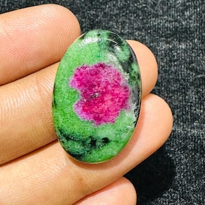 Puede incluir: Una piedra preciosa en forma de cabujón ovalado con un centro rosa magenta vibrante rodeado por una matriz de zoisita verde y negra moteada. La piedra tiene una superficie lisa y pulida con un acabado brillante y variaciones de color.
