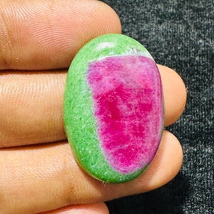 Può includere: Una gemma a cabochon di forma ovale caratterizzata da un centro rosa magenta vibrante circondato da un bordo verde lime brillante. La pietra presenta una superficie liscia e lucida con un netto contrasto cromatico tra il centro e il bordo.