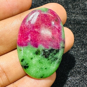 May include: An oval-shaped polished gemstone featuring a vibrant magenta-pink section at the top and a bright lime-green section at the bottom, with scattered dark black mineral inclusions throughout the green area and a smooth, reflective surface.