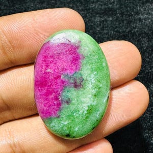 Può includere: Una pietra preziosa lucidata di forma ovale che presenta un mix vibrante di colori magenta intenso e verde brillante. La pietra ha una superficie liscia e lucida con variazioni di colore naturali e inclusioni visibili nel cabochon.