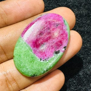 Può includere: Una gemma cabochon di forma ovale caratterizzata da un vibrante centro rosa magenta circondato da un bordo verde lime brillante. La pietra presenta una superficie liscia e lucida con variazioni di colore naturali e piccole inclusioni scure.
