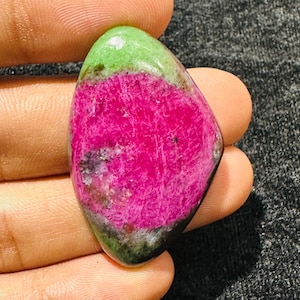 May include: A polished, teardrop-shaped ruby zoisite gemstone featuring a vibrant, saturated magenta-pink center surrounded by a smooth, light green border with dark mineral inclusions, showcasing a natural, glossy, and multi-colored surface.