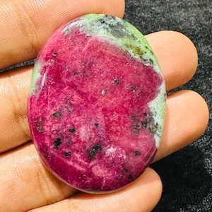 Op de afbeelding: Een ovaalvormige gepolijste robijn-zoisiet edelsteen met een levendig magenta-roze centrum met donkere zwarte mineraalinsluitingen en lichtgroene randen, met een gladde, glanzende oppervlaktestructuur over de gehele steen.