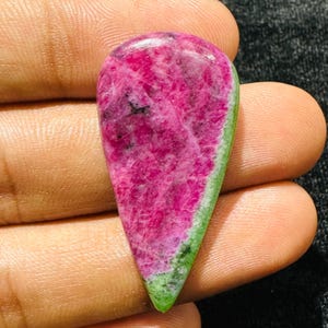 Peut inclure: Une pierre précieuse cabochon en forme de goutte avec un centre magenta vif aux textures cristallines naturelles et inclusions sombres, bordée par une bande distincte de minéral vert forêt opaque sur le bord droit et la pointe inférieure.