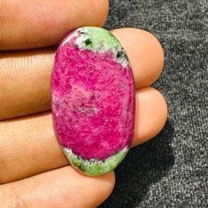 Può includere: Una pietra preziosa cabochon di forma ovale caratterizzata da un centro rosa magenta vibrante circondato da inclusioni minerali screziate di verde chiaro e verde scuro. La pietra presenta una superficie liscia e lucida con una finitura brillante e variazioni di colore naturali.