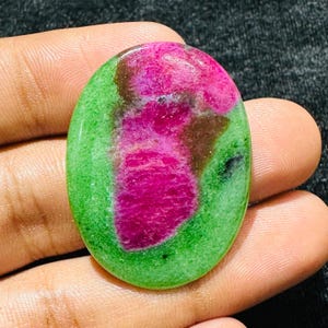 以下が含まれることがあります： 鮮やかな緑色のベースカラーに、深いマゼンタ色と濃い茶色の不規則な斑点が表面全体に広がる、楕円形に研磨された宝石。滑らかで光沢があり、光を反射する仕上げが施されています。