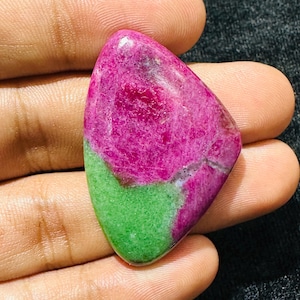 Può includere: Una gemma di rubino zoisite lucidata a forma di goccia, caratterizzata da un contrasto vibrante tra inclusioni di rubino magenta intenso e una base in zoisite verde brillante, con una superficie liscia e texture minerali naturali.