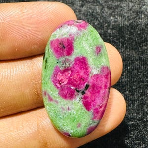 May include: An oval-shaped polished gemstone featuring a vibrant mix of deep magenta-pink ruby inclusions embedded within a light green zoisite matrix, displaying a smooth, reflective surface with natural colour variations throughout the stone.