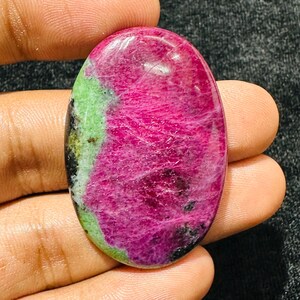 May include: An oval-shaped cabochon gemstone featuring a vibrant magenta-pink center with natural inclusions, bordered by a contrasting light green mineral section and small patches of black, polished to a smooth, glossy finish.