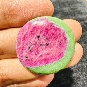 May include: A circular polished gemstone featuring a vibrant magenta centre with small black inclusions, surrounded by a smooth, light green border. The stone has a flat, rounded shape with a glossy finish, displaying natural mineral textures.