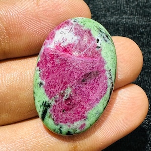 Pode incluir: Uma pedra preciosa cabochão de formato oval com um centro rosa magenta vibrante rodeado por uma borda verde clara com inclusões minerais pretas dispersas. A pedra tem uma superfície lisa e polida com um acabamento brilhante.
