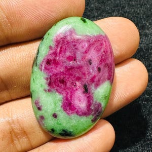 Puede incluir: Una piedra preciosa cabujón de forma ovalada que presenta un centro rosa magenta vibrante rodeado por una matriz verde claro con inclusiones minerales negras dispersas. La piedra pulida tiene una superficie lisa y redondeada y un acabado brillante.