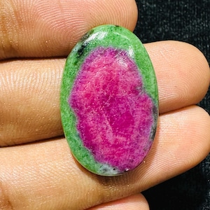 May include: An oval-shaped cabochon gemstone featuring a vibrant magenta-pink centre surrounded by a distinct border of opaque forest green material. The stone has a smooth, polished surface with visible natural inclusions and colour variations.