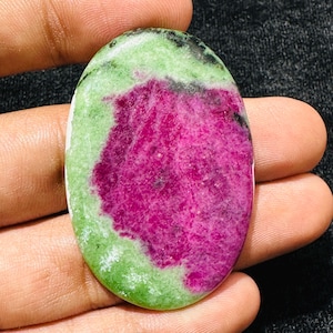 Può includere: Una pietra preziosa a cabochon di forma ovale caratterizzata da un centro rosa magenta vibrante circondato da un bordo verde chiaro con piccole inclusioni nere. La pietra presenta una superficie liscia e lucida con un netto contrasto cromatico.