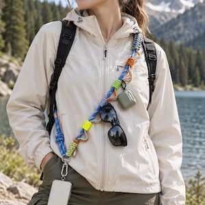May include: A blue rope crossbody phone lanyard featuring multi-coloured paracord accents in neon yellow and orange. The lanyard includes a metal clip attached to a smartphone, a pair of black sunglasses, and a sage green earbud case.