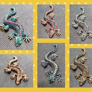 Puede incluir: Seis broches de lagarto con joyas de diferentes colores. Los lagartos están todos en la misma pose, con la cola enroscada y la cabeza inclinada hacia un lado. Las broches están hechas de metal y están decoradas con diamantes de imitación. Los colores de las broches son azul, rosa, plata, oro, azul verdoso y negro.
